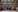 A girl sat at a desk in the library at her laptop, looking at a book. The background is shelves of books. 