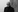 A black and white image of a man with glasses standing next to a wall looking into the carmera. 