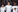 Three rugby players wearing an England kit, stand in a line with their arms around each other, singing. It is raining in the picture. 