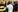 Two women are in the shot, one whose face we can see and the other faces away from the camera. The woman whose face is visible is meeting the other woman and smiling broadly.